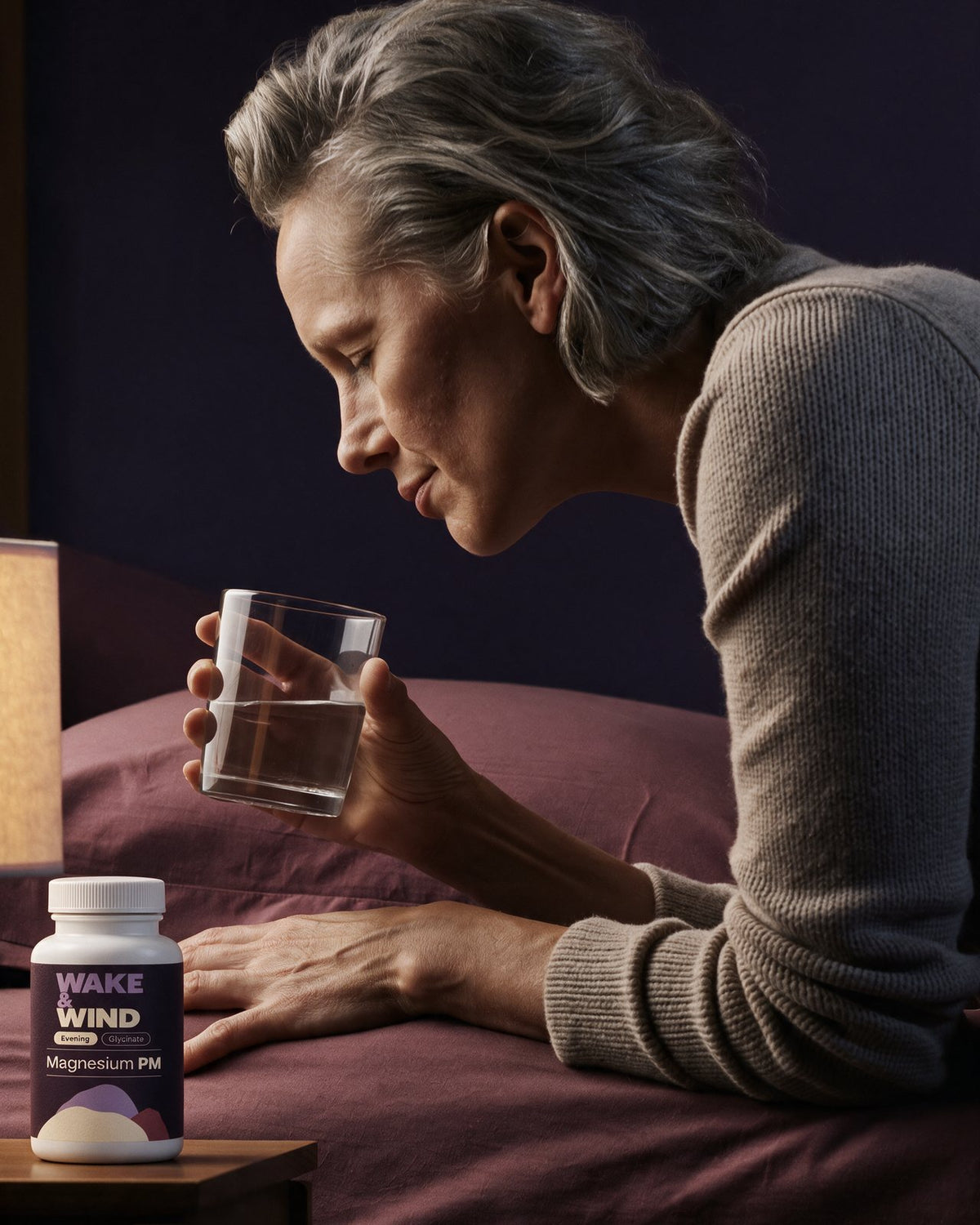 A woman in her late fifties drinking water by a low lamp at 21:50, Magnesium PM on the bedside table