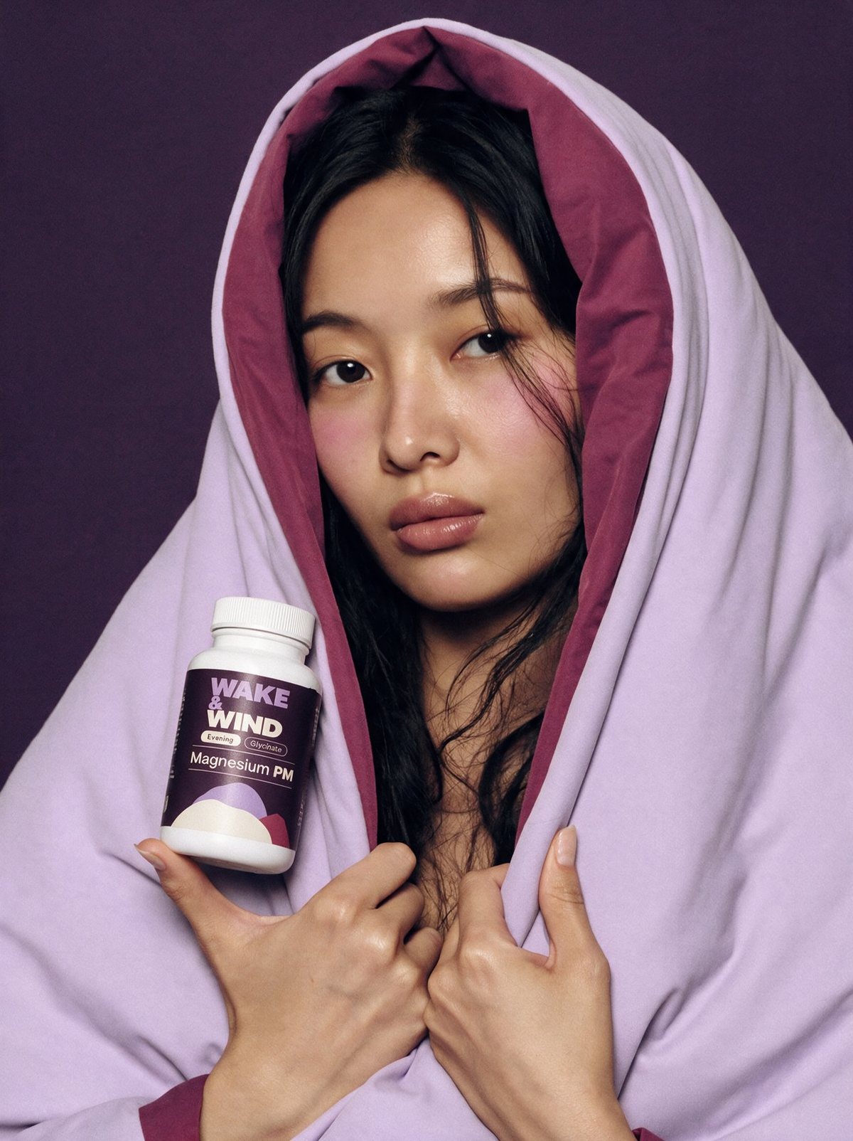 A woman wrapped in a lilac duvet holding a Magnesium PM magnesium glycinate bottle in the evening