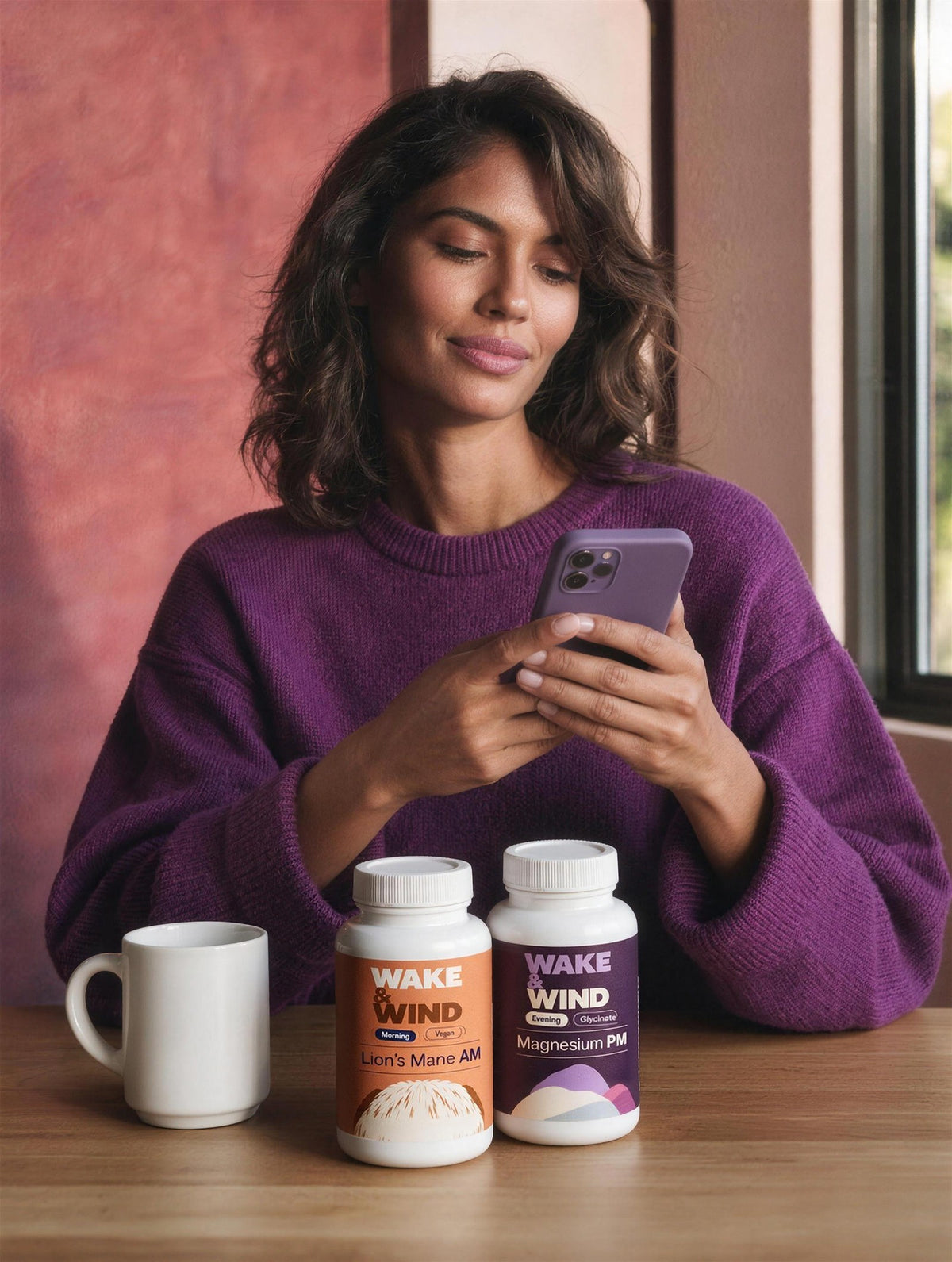 A woman using the Wake & Wind app on her phone at a table with Lion's Mane AM and Magnesium PM bottles